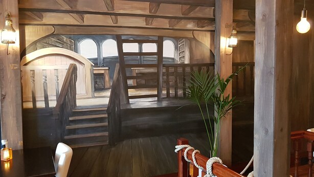 Een houten interieur met een trap die naar een hoger gelegen verdieping leidt, een kamerplant en uitzicht op buiten.