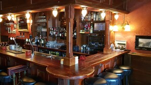 Een bar met een lange toonbank, meerdere krukken en een goed gevulde plank met diverse flessen en glazen.