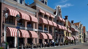 Een rij bakstenen gebouwen met rode en witte streepbeschutte zonneschermen, een bord met 'Hotel Restaurant Anna Caspari', zitplaatsen buiten en een heldere blauwe lucht.