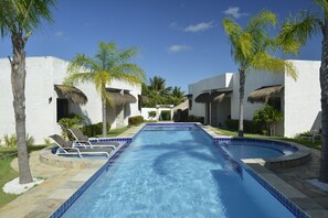 A swimming pool with lounge chairs and palm trees, surrounded by white buildings with thatched roofs. - Oasis Tajaja (Conde)