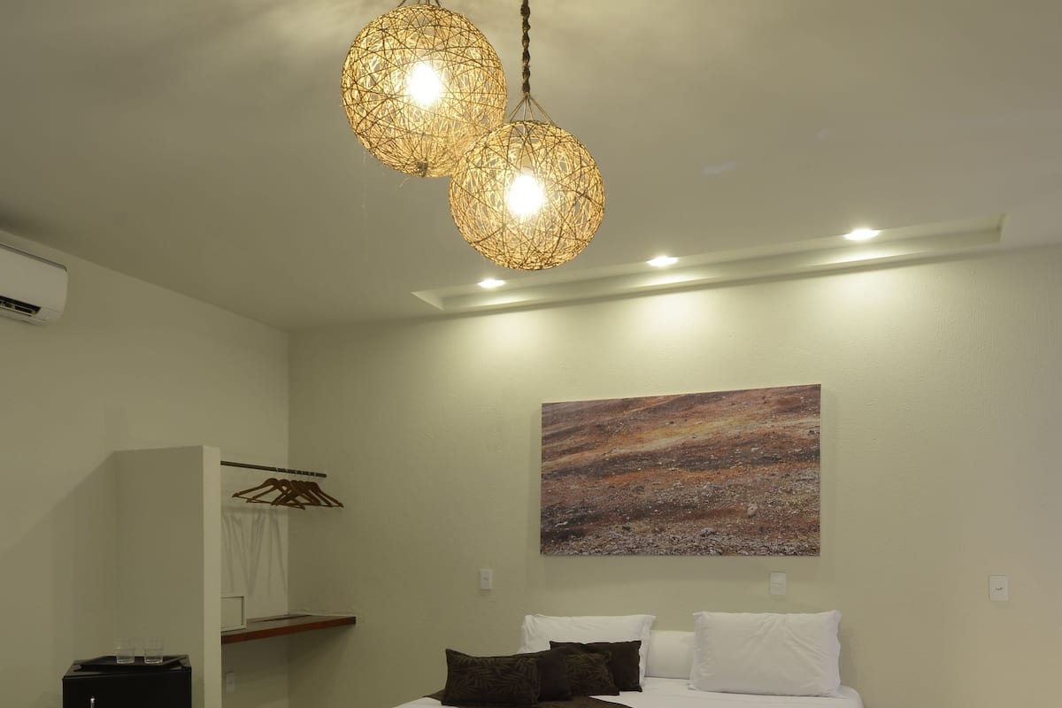 Quarto moderno com uma cama grande, um banquinho de madeira, um minibar preto e uma luminária esférica singular.