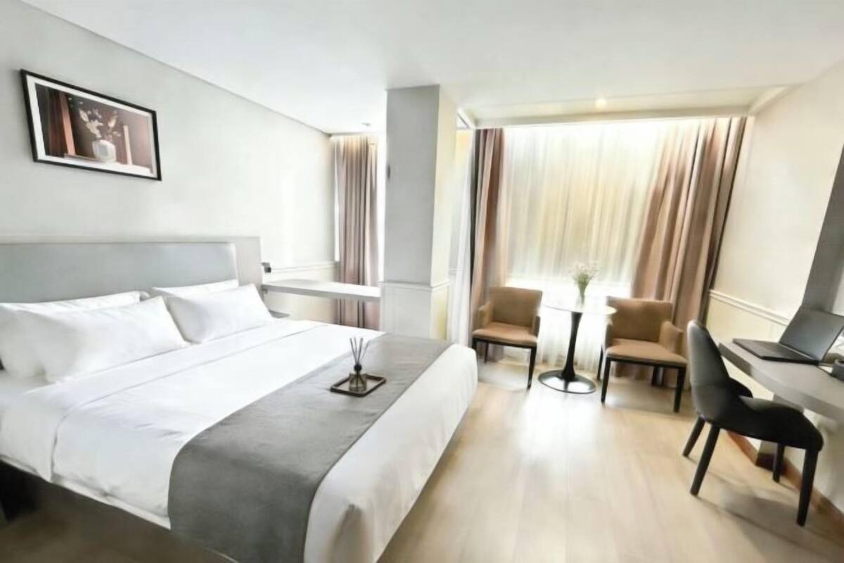 Ein Hotelzimmer mit Bett, Schreibtisch, zwei Stühlen, einem kleinen Tisch und einem Fenster mit Vorhängen.