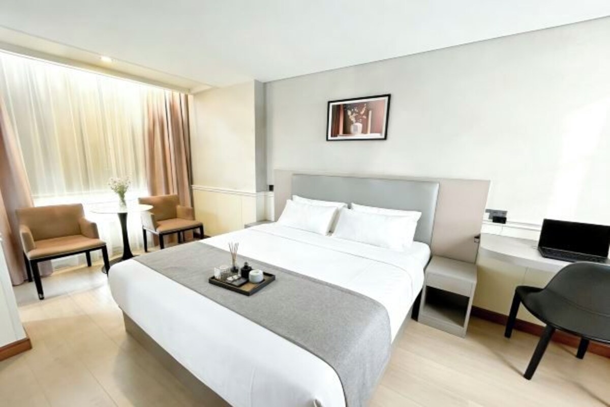 A hotel room with a bed, two chairs, a desk with a laptop, a small table with a vase, and a framed picture on the wall.