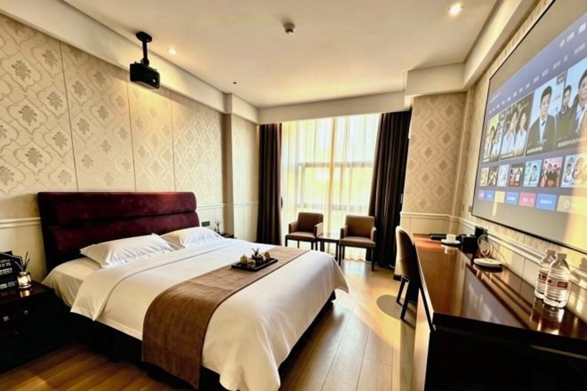 Une chambre d’hôtel avec un grand lit, un bureau équipé d’un ordinateur et un écran affichant plusieurs images.