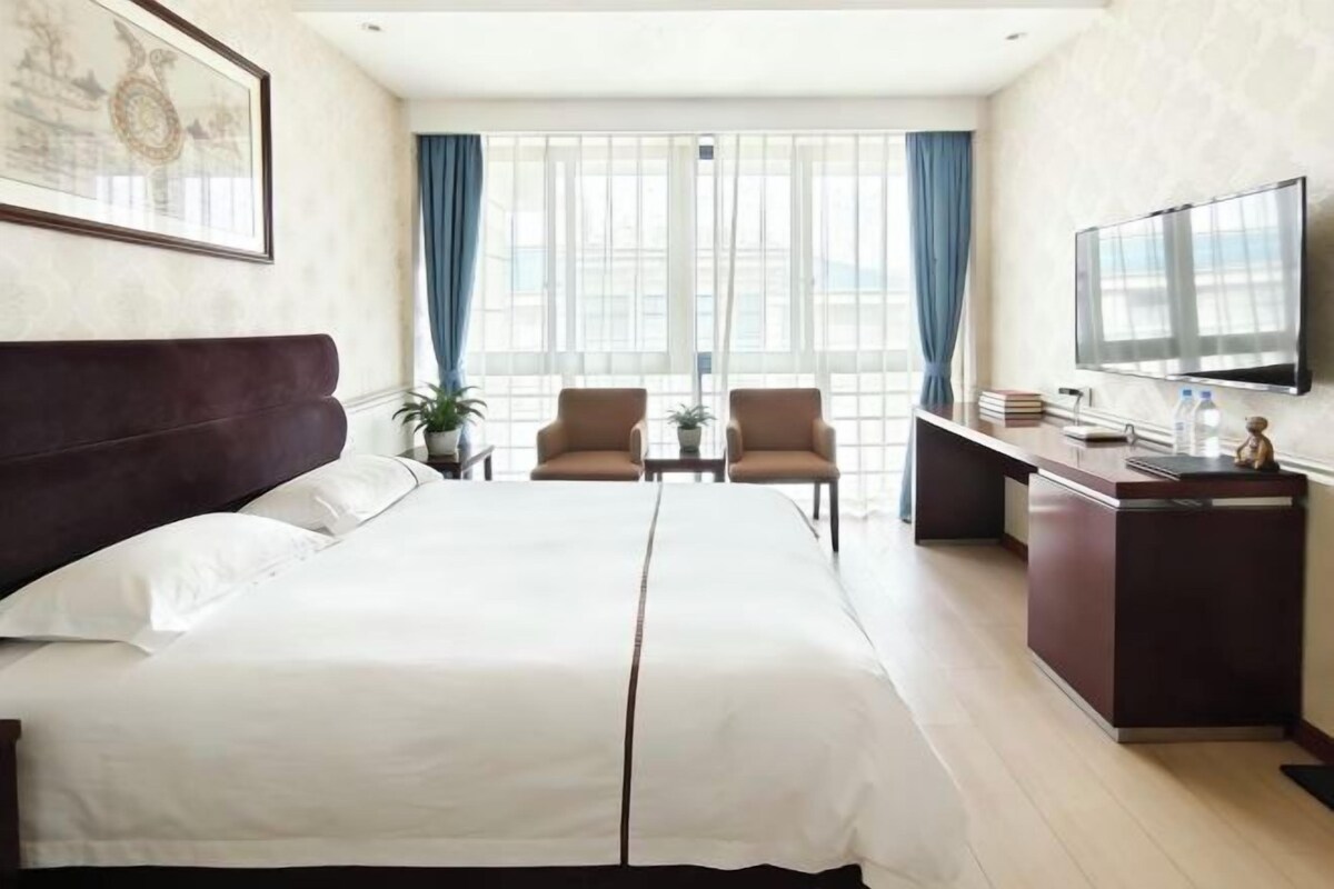 Ein Hotelzimmer mit einem Bett, zwei Sesseln, einem Schreibtisch, einem Fernseher und einem Fenster mit Vorhängen.