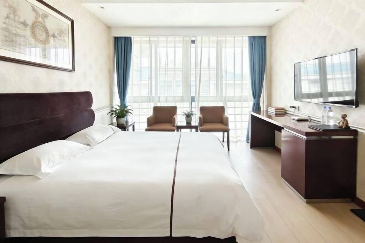 Ein Hotelzimmer mit einem Bett, zwei Sesseln, einem Schreibtisch, einem Fernseher und einem Fenster mit Vorhängen.