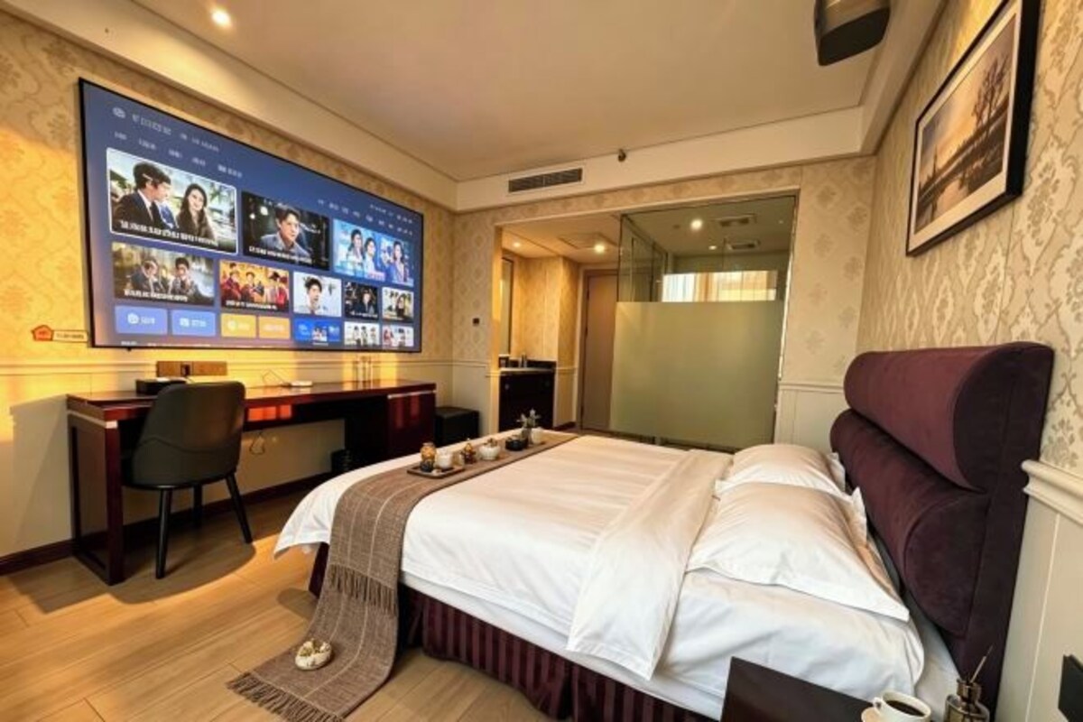 A hotel room with a large flat-screen TV mounted on the wall displaying multiple video feeds, a bed with white linens, a desk with a chair, and a small table with a cup and a bottle.