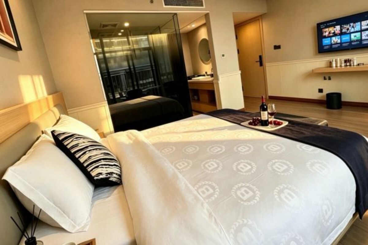 A hotel room with a bed, a TV, a small table with a bottle and glasses, and a door leading to a balcony.