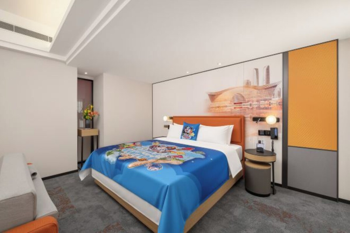A hotel room with a large bed, a nightstand, a wall-mounted lamp, and a cityscape artwork on the wall.