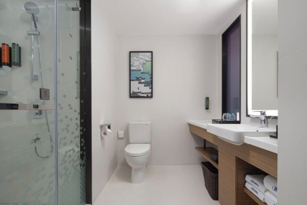 A modern bathroom with a glass shower enclosure, a white toilet, a wooden vanity with a mirror, and a framed artwork on the wall.