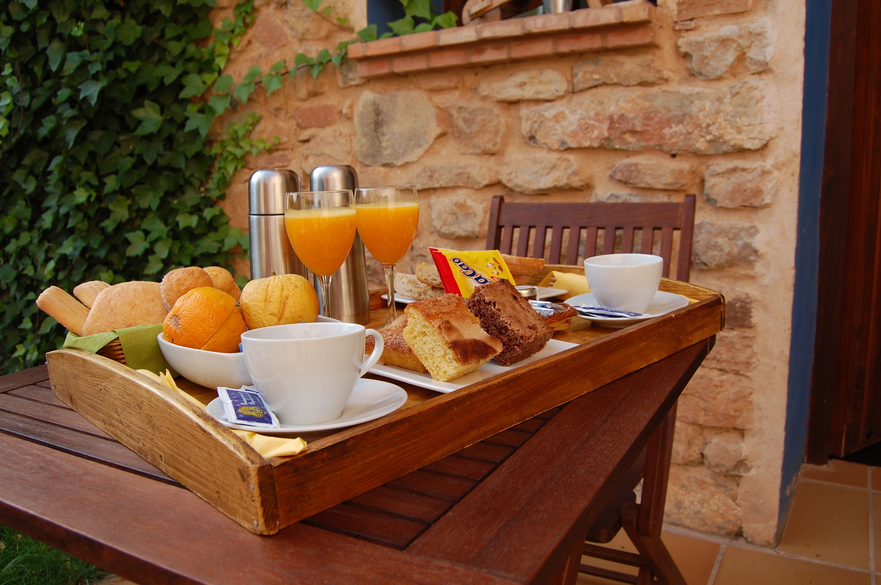 Una mesa de madera con una bandeja que contiene pan, naranjas, una taza, un vaso de jugo de naranja y una bolsita de mantequilla.