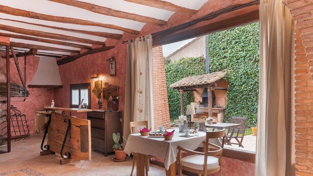 Un comedor rústico con una mesa dispuesta para dos, sillas de madera y vistas a un muro de ladrillo y follaje exterior.