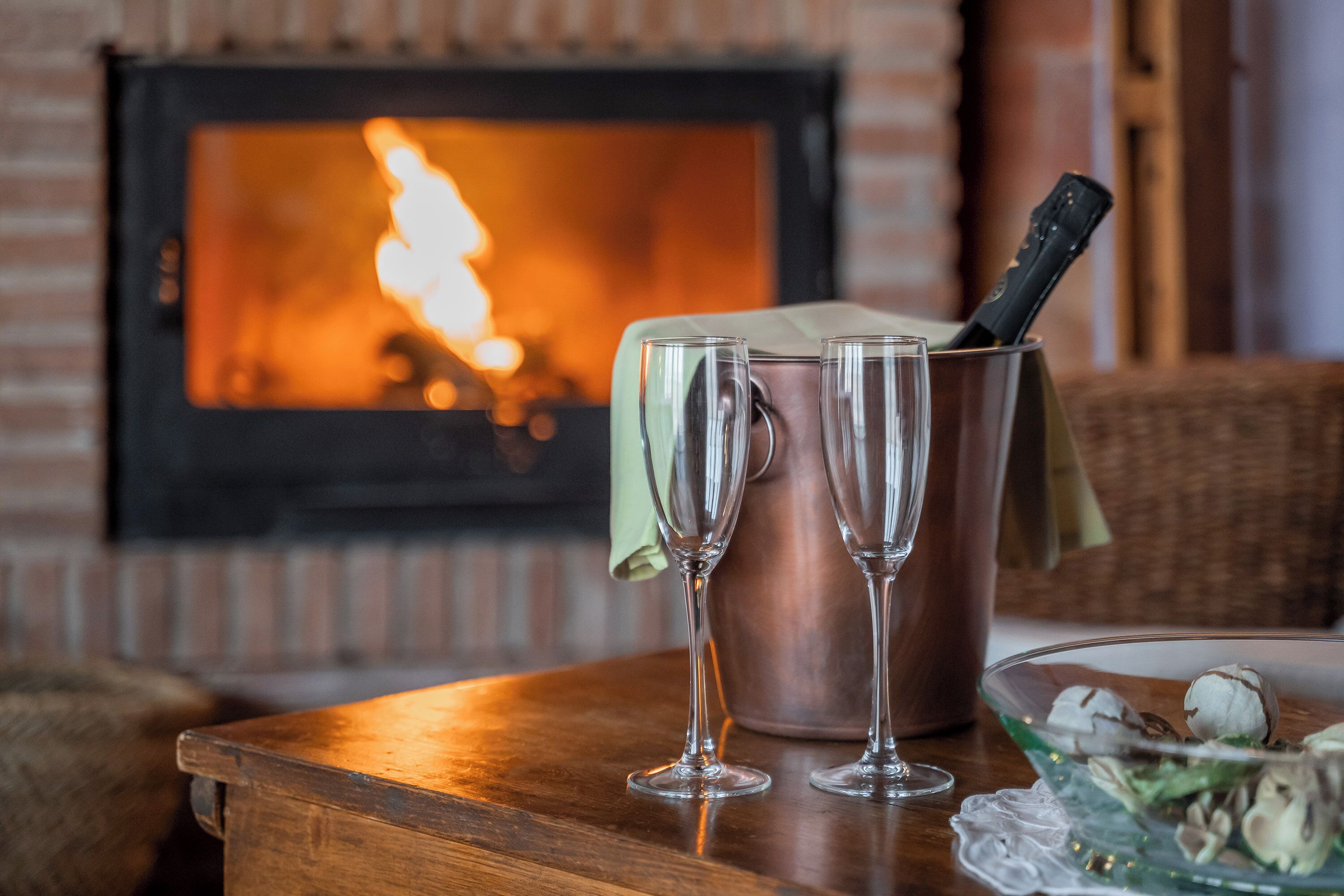 Ein Holztisch mit zwei Sektgläsern, einem Kupferkübel mit Flaschenöffner und einer Glaskugel mit Nüssen. Im Hintergrund ist ein Kamin mit brennendem Feuer zu sehen.