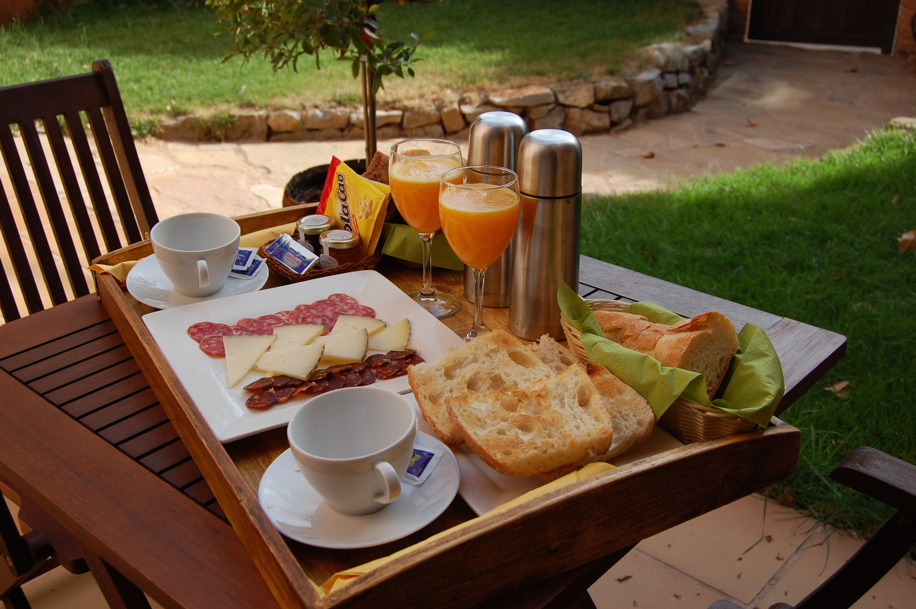 A breakfast tray with cheese, ham, bread, and juice on a wooden table outdoors.