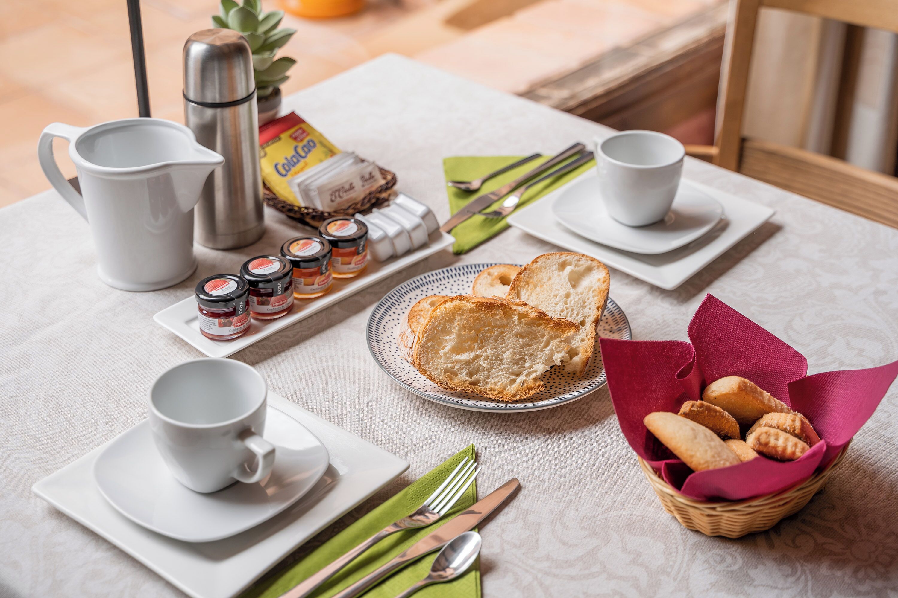 Een ontbijttafel met een karaf, kopjes, een bord met brood en een mand met gebak.