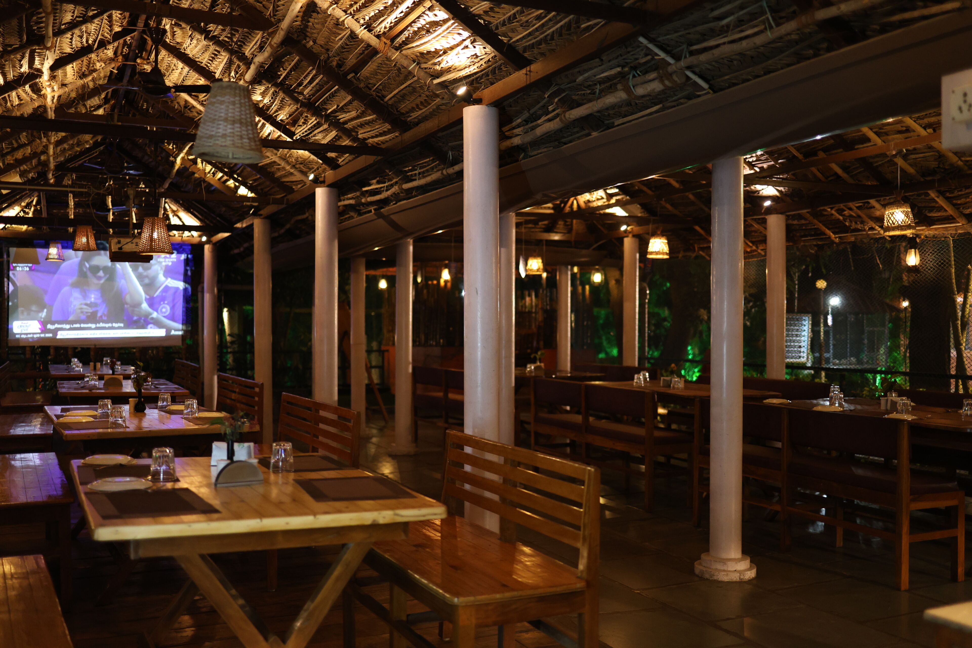 An indoor dining area with wooden tables and chairs, a thatched roof ceiling, and hanging light fixtures.