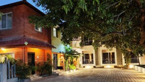 Un patio con un sendero empedrado, un edificio de fachada roja y un árbol que da sombra.