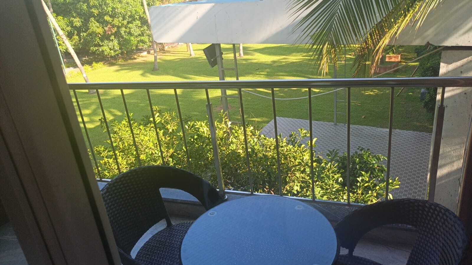 Uma varanda com duas cadeiras de vime e uma mesa pequena, com vista para um jardim bem cuidado com gramado e palmeiras.