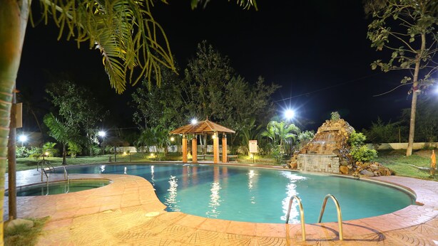 Una piscina nocturna con un pabellón y una estructura de piedra, rodeada de árboles e iluminada por farolas.