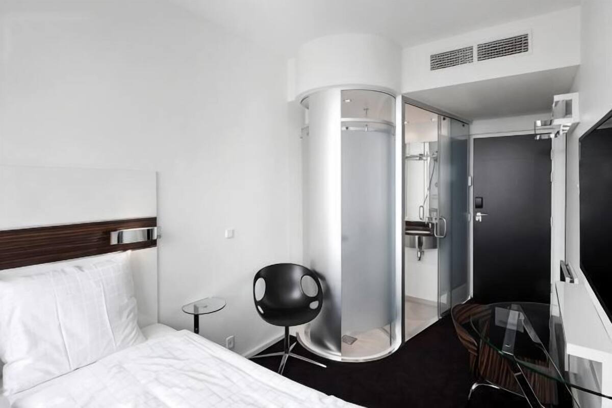 Una habitación de hotel con una cama, cabina de ducha de vidrio, una mesita y una silla.