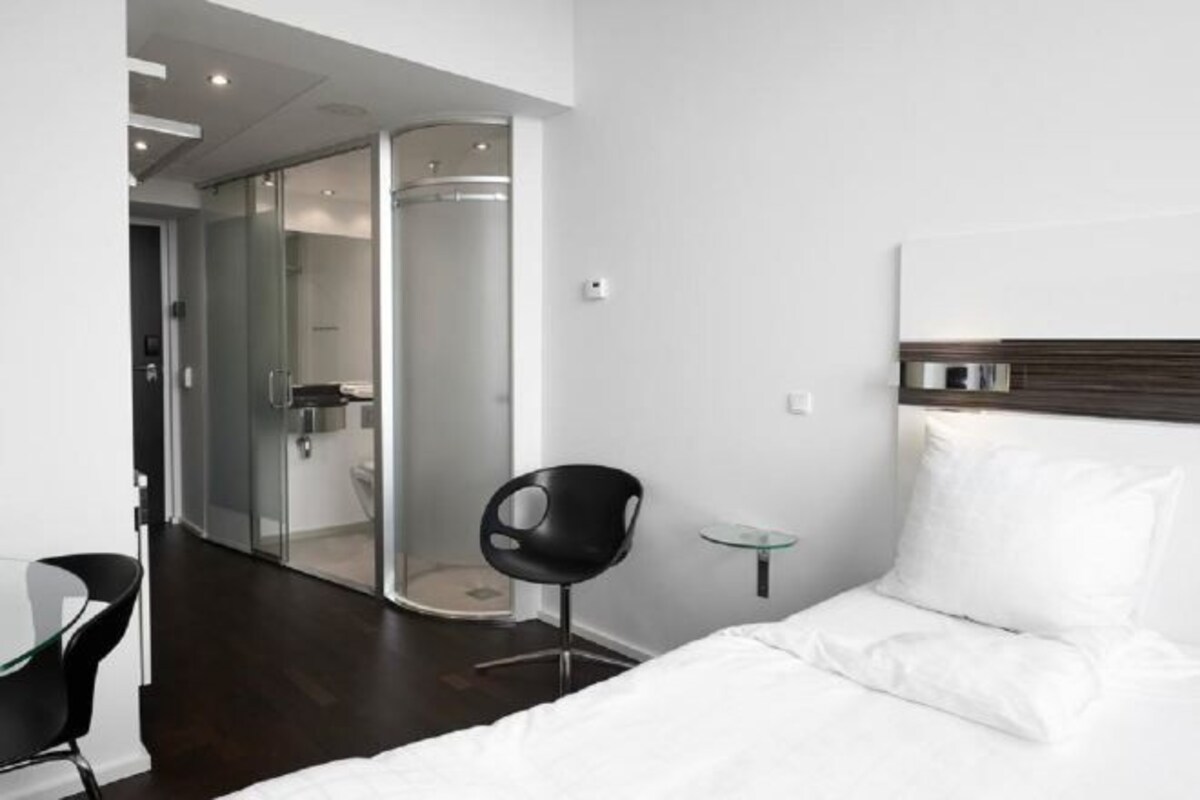 Een moderne hotelkamer met een bed, een douchecabine van glas, een zwarte stoel en een glazen tafeltje.