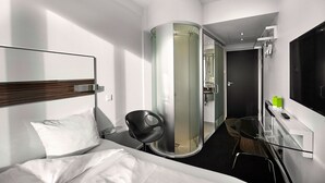 Ett modernt hotellrum med en säng, en duschkabin med glasväggar, ett litet bord med en stol och en tv på väggen.