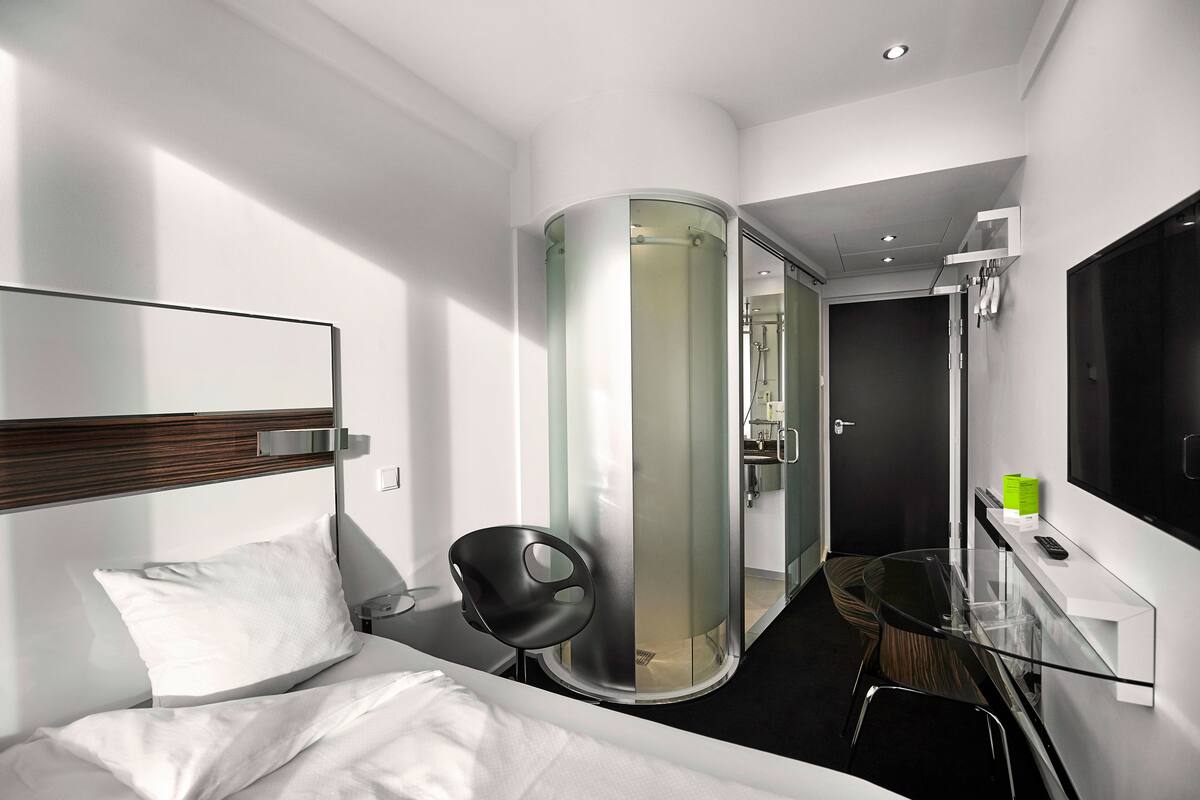 Una habitación de hotel moderna con una cama, ducha envuelta en vidrio, una mesita con una silla y un televisor en la pared.