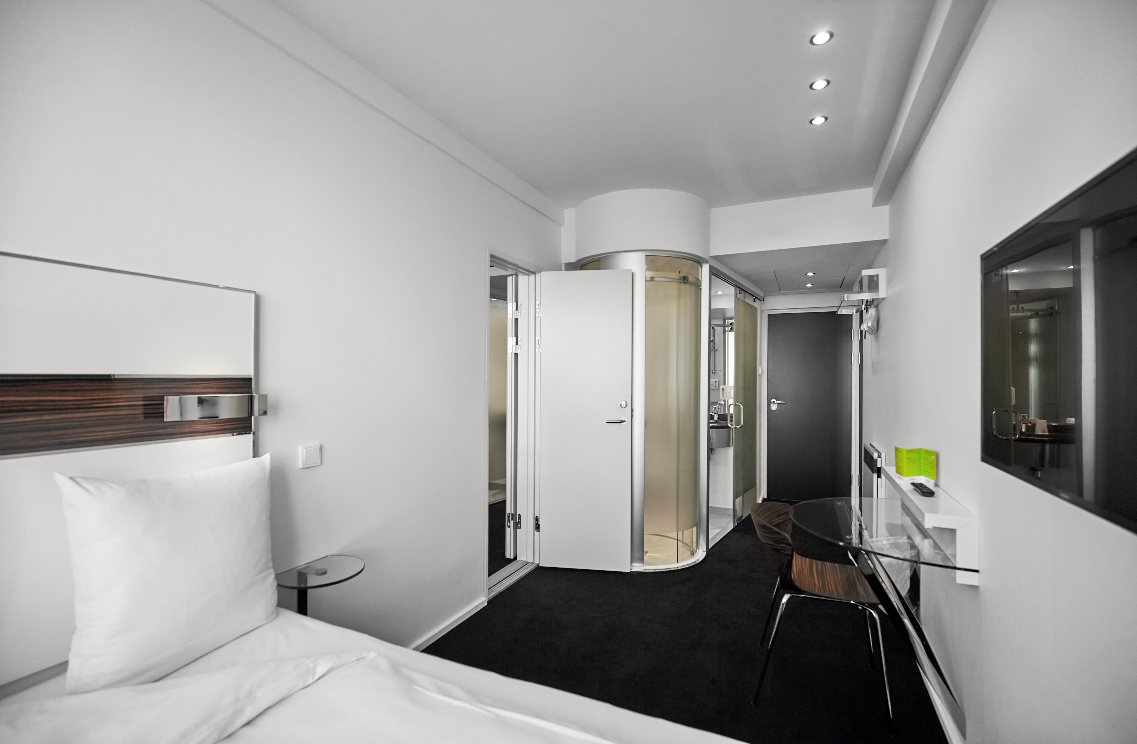 Una habitación de hotel moderna con una cama, una mesita redonda pequeña, un separador de vidrio y una zona de ducha.
