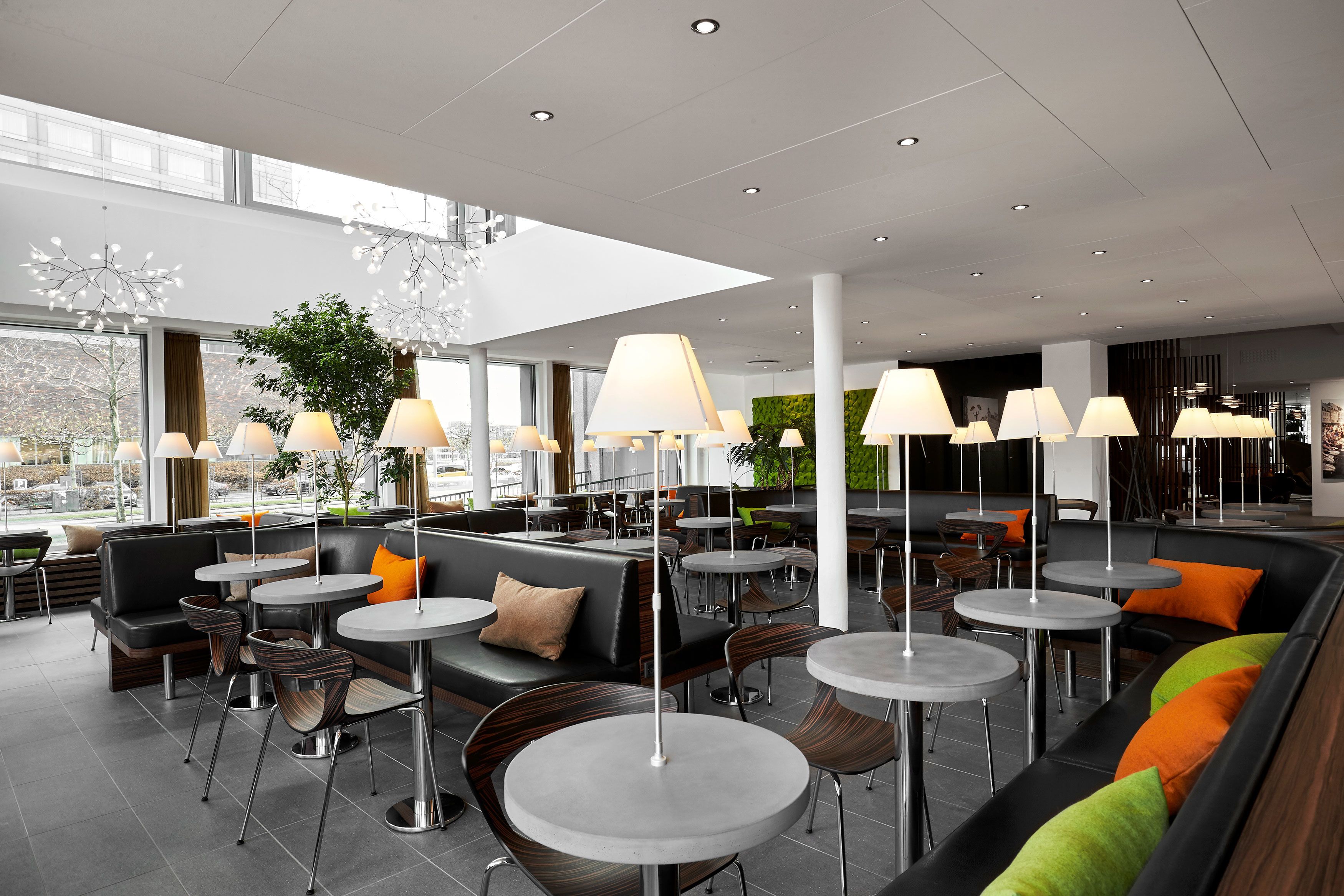 A modern indoor seating area with round tables and sofas, overhead lighting, and large windows.