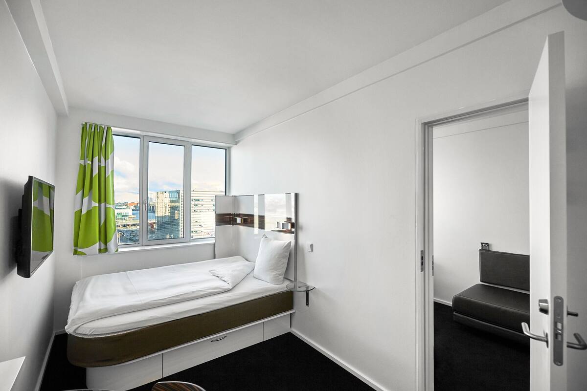 Habitación de hotel compacta con una cama individual, una ventana con cortinas, una mesita de noche, un espejo y una puerta que da a una zona de estar.