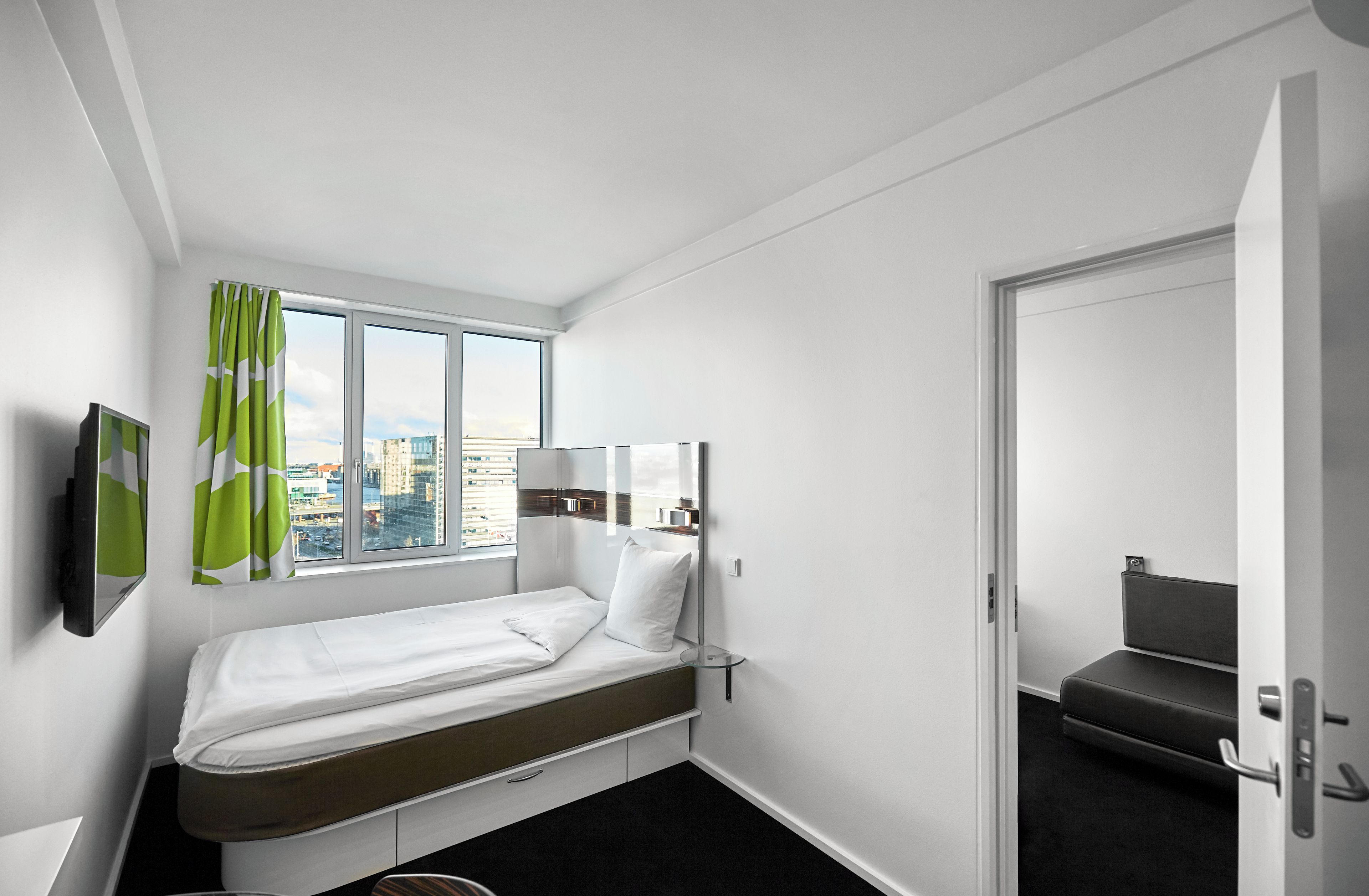 Une chambre d’hôtel avec un lit simple, une fenêtre avec des rideaux, un téléviseur à écran plat et une porte donnant sur un coin salon.