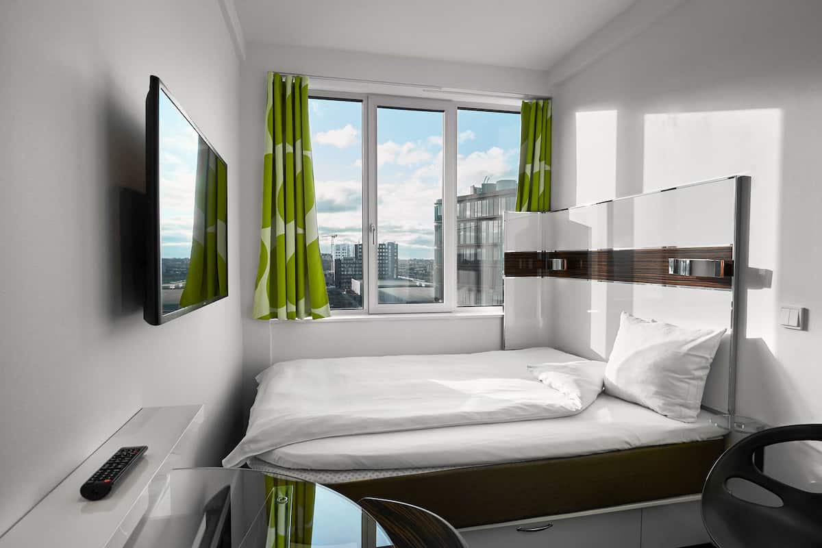 Una habitación de hotel moderna con una cama individual, un televisor montado en la pared, un escritorio con un control remoto y una ventana con cortinas.