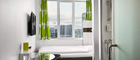 A compact hotel room with a bed, a desk with a chair, a window with curtains, a television mounted on the wall, and a door with a handle.