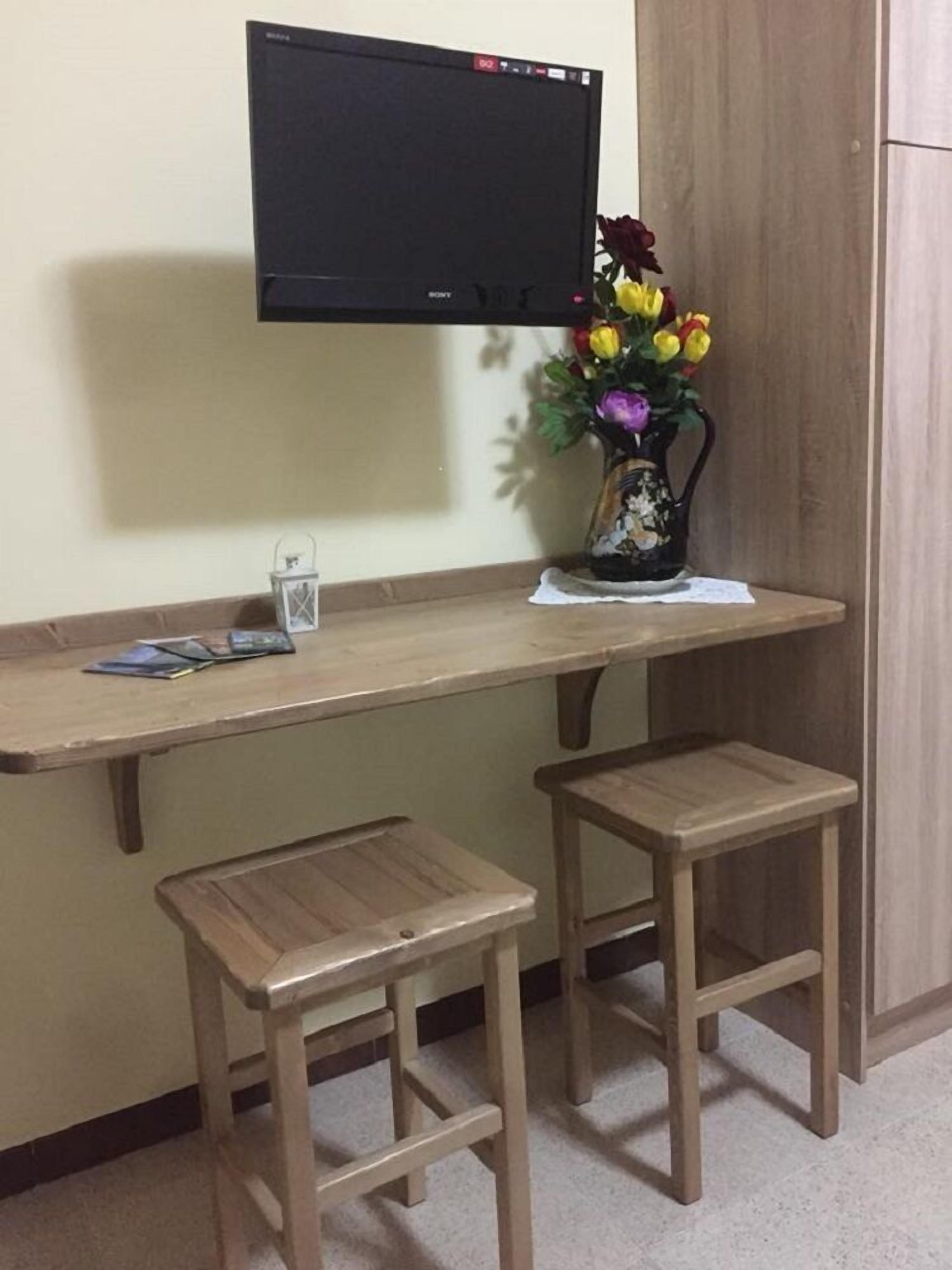 A small workspace with a wooden desk, two stools, a television mounted on the wall, and a vase with flowers.