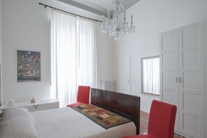 Une chambre à coucher avec un lit, un lustre, une fenêtre avec des rideaux, un radiateur et une armoire.