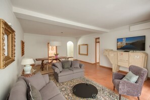 Sala de estar com sofá, poltrona e televisão. Há uma mesa de jantar com cadeiras e um abajur. É visível um armário com adega.