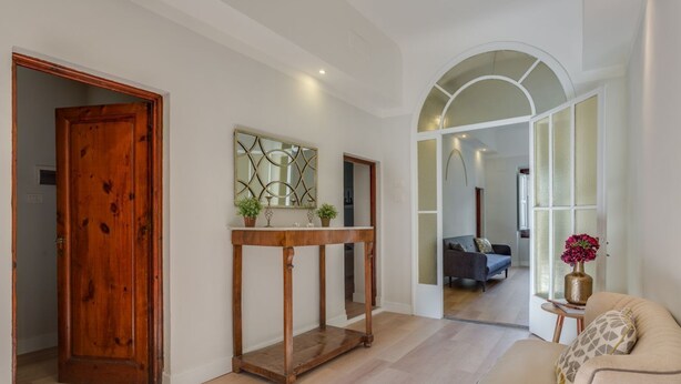 A spacious hallway with a wooden door, a console table with plants, a sofa, and a mirror reflecting a living room.