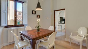A dining area with a wooden table, white chairs, a pendant light, and a view of the kitchen through an open window.