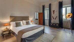 A bedroom with a large bed, a nightstand with a lamp, a dresser with a mirror, a window with curtains, and a wall-mounted radiator.