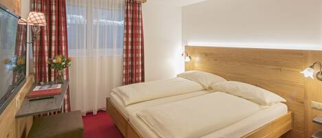 Habitación de hotel con una cama de madera, un escritorio con lámpara, un televisor y una ventana con cortinas.