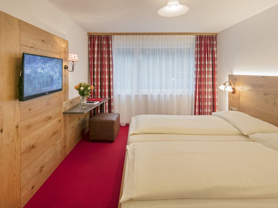 Ein Hotelzimmer mit Holz-Kopfteil, einem an der Wand befestigten Flachbildfernseher, einem kleinen Tisch mit Blumenvase, einer Bank und einem großen Bett mit weißer Bettwäsche.