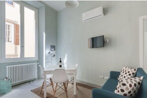 A room with a white table, a blue sofa, a TV mounted on the wall, and a window with shutters. - Apartments Florence - Taddea Terrace (Firenze)