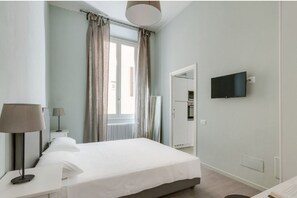 A hotel room with a bed, a TV mounted on the wall, a window with curtains, a bedside table with a lamp, and a radiator.