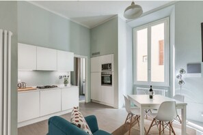 Une cuisine moderne avec des meubles blancs, un coin repas doté d’une table et de chaises blanches, et une grande fenêtre qui laisse entrer la lumière naturelle.