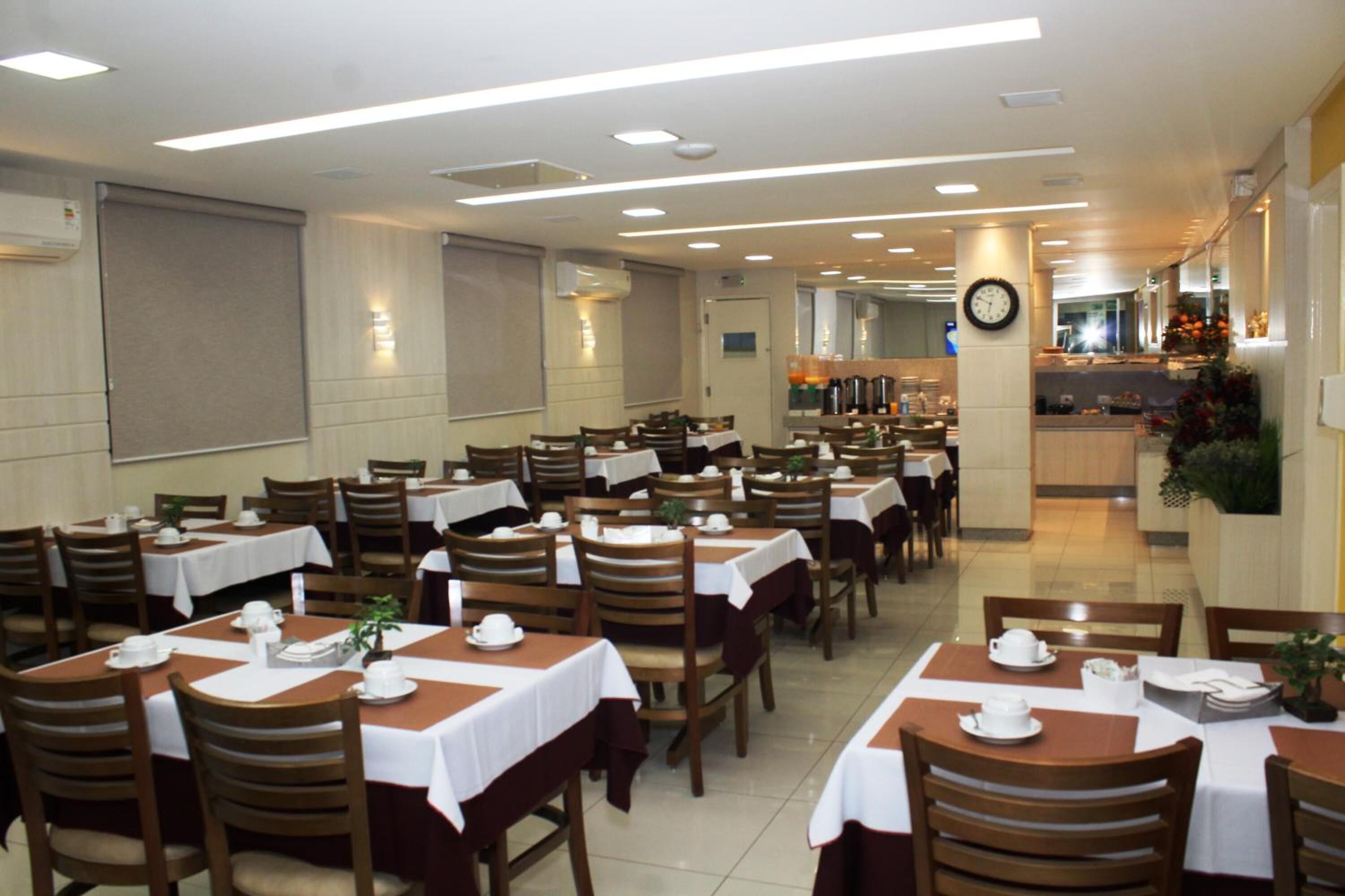 A dining area with multiple tables and chairs, some with white tablecloths and others with brown covers. There are ceiling lights and a clock on the wall.