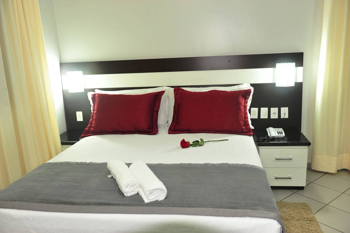 A neatly made bed with red and white pillows, a rolled white towel, and a single red rose.