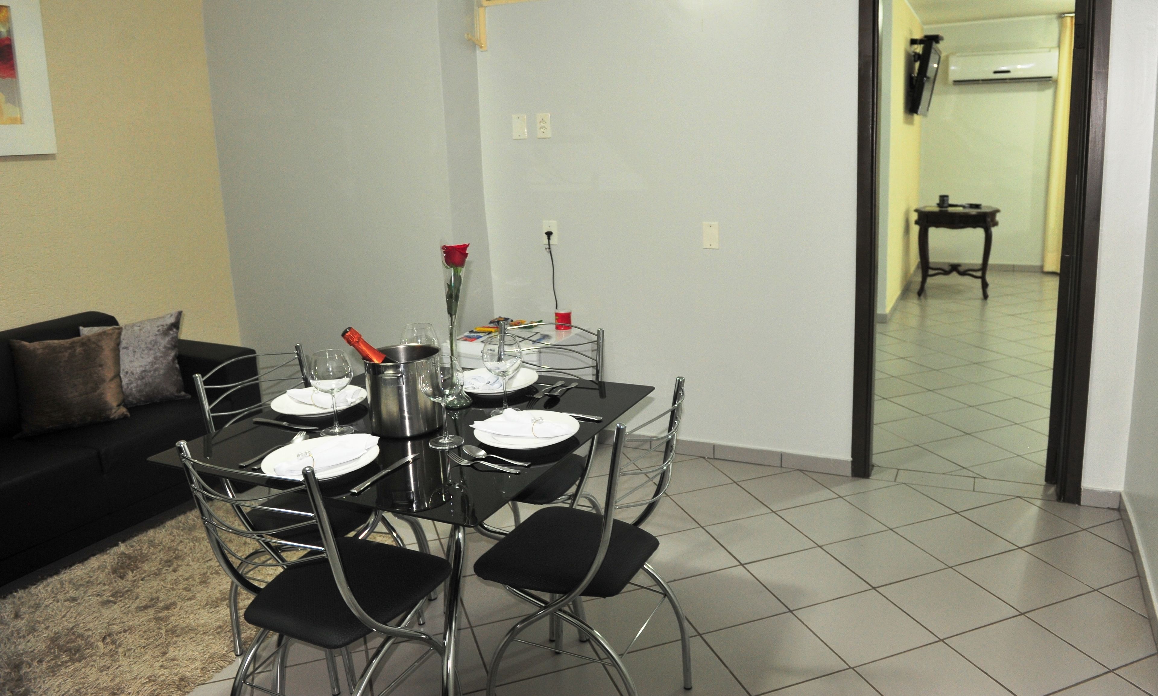 Un comedor con una mesa dispuesta para cuatro, incluyendo copas de vino y una botella de champán. Se observa un sofá negro con cojines al fondo.