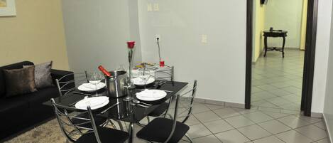 Un comedor con una mesa dispuesta para cuatro, incluyendo copas de vino y una botella de champán. Se observa un sofá negro con cojines al fondo.