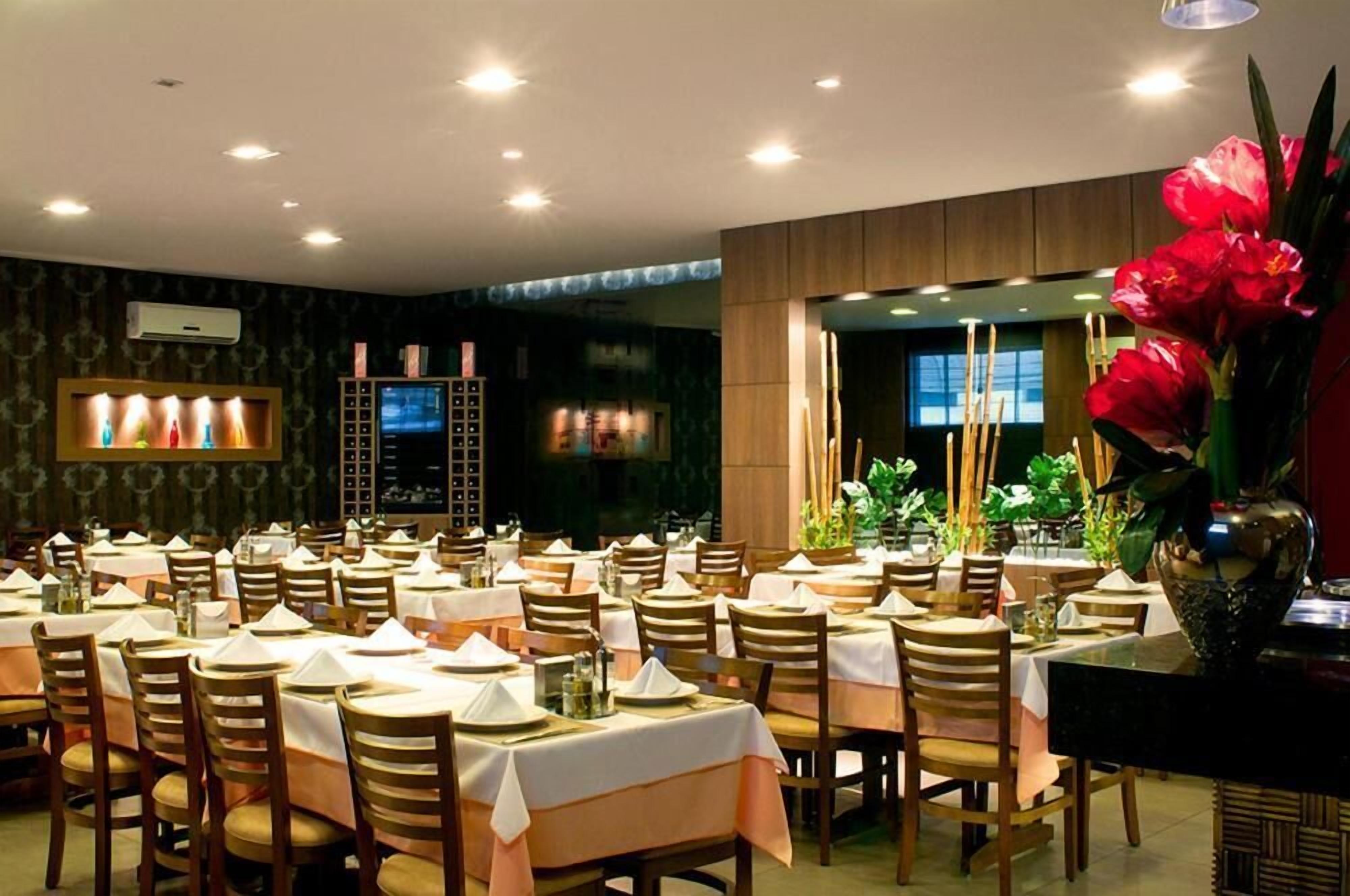 A restaurant with multiple tables set for dining, a large decorative vase with flowers, and a television screen on the wall.