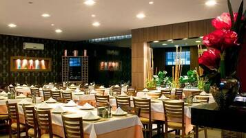 A restaurant with multiple tables set for dining, a large decorative vase with flowers, and a television screen on the wall.